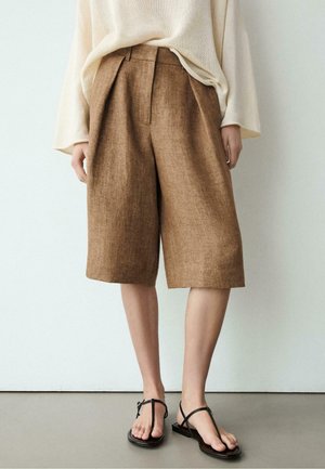 Braune knielange, weit geschnittene Shorts, getragen mit schwarzen Riemchensandalen und einem lockeren cremefarbenen Stricktop, vor einem schlichten weißen Hintergrund stehend.