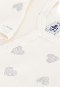 Body per neonato color crema realizzato in cotone morbido, presenta motivi a cuore glitterato argento, con bottoni a pressione e un'etichetta sul collo.