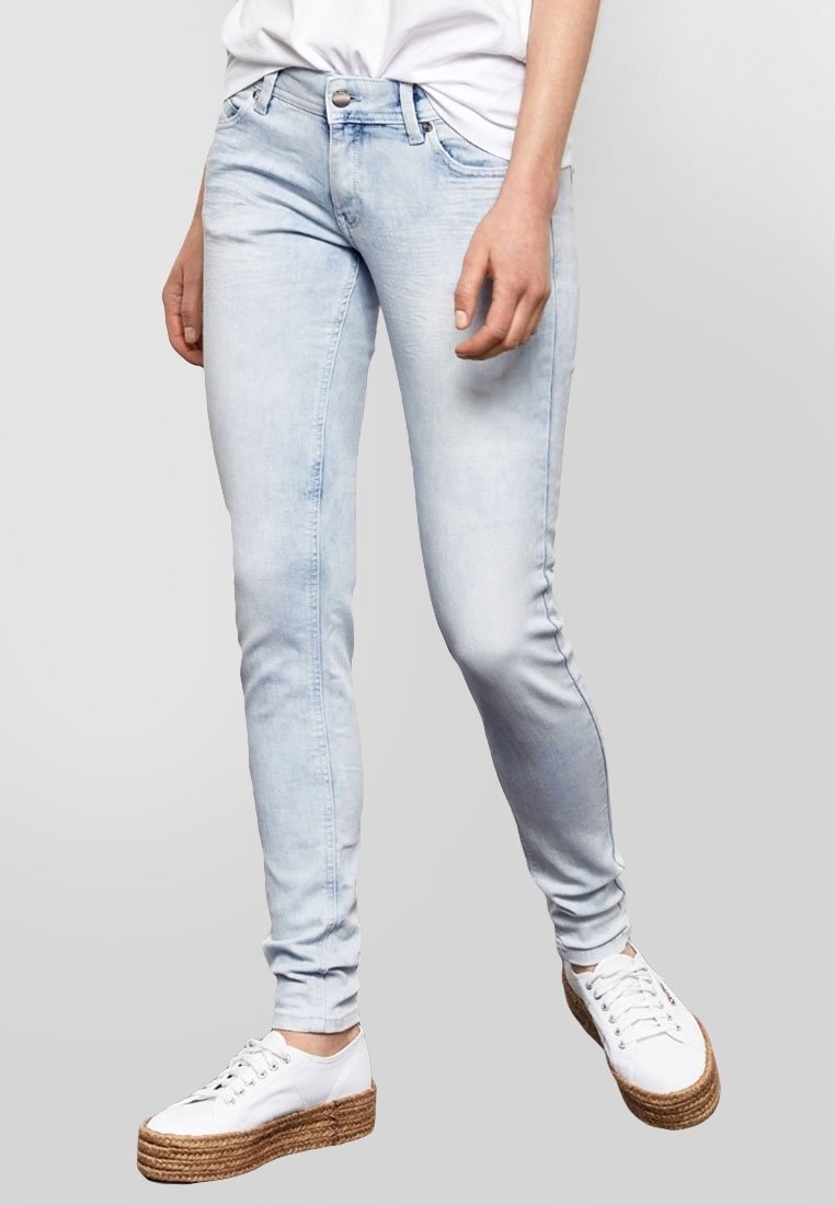 Jean skinny bleu clair en denim, avec un délavage moyen, un léger effet de dégradé, cinq poches et une fermeture à bouton. Associé à des baskets plateformes blanches.