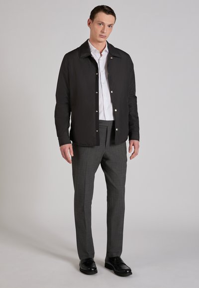 Chaqueta ligera negra con cierre de botones a presión en la parte delantera, combinada con pantalones grises a cuadros y zapatos negros. Diseño limpio y ajuste entallado.