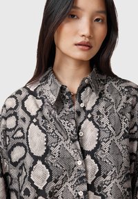 Camicia con bottoni caratterizzata da una stampa di serpente nera e crema, con colletto e tessuto testurizzato. I bottoni frontali sono visibili.
