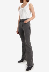 Haut débardeur blanc à volants avec détail à nouer, associé à un jean évasé gris foncé. Le mannequin porte des bottines noires.