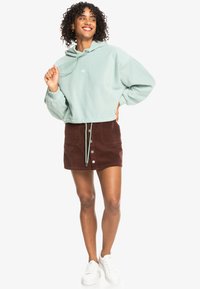 Sudadera corta verde claro con cordón, combinada con una minifalda de pana marrón con botones frontales, llevada con zapatillas blancas.
