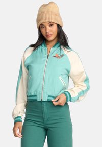 Light teal bomber jacket with cream sleeves, embroidered detail, ribbed collar, and cuffs. Paired with green pants. Textured knit beanie.