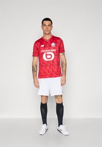 Rød fodboldtrøje med et tekstureret mønster, hvide shorts, sorte stribede sokker og hvide fodboldstøvler. Har holdlogo og sponsorbranding.