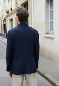 Blazer marine en tissu texturé, doté d'un col cranté, d'une fente simple à l'arrière et de boutons marron sur les manches, associé à un pantalon clair.
