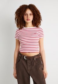 Νεαρή γυναίκα με κατσικίσιο μαλλί, φορώντας λευκό και κόκκινο ριγέ κοντό τοπ και καφέ ψηλόμεσο παντελόνι με ζώνη, στέκεται μπροστά σε λευκό τοίχο.