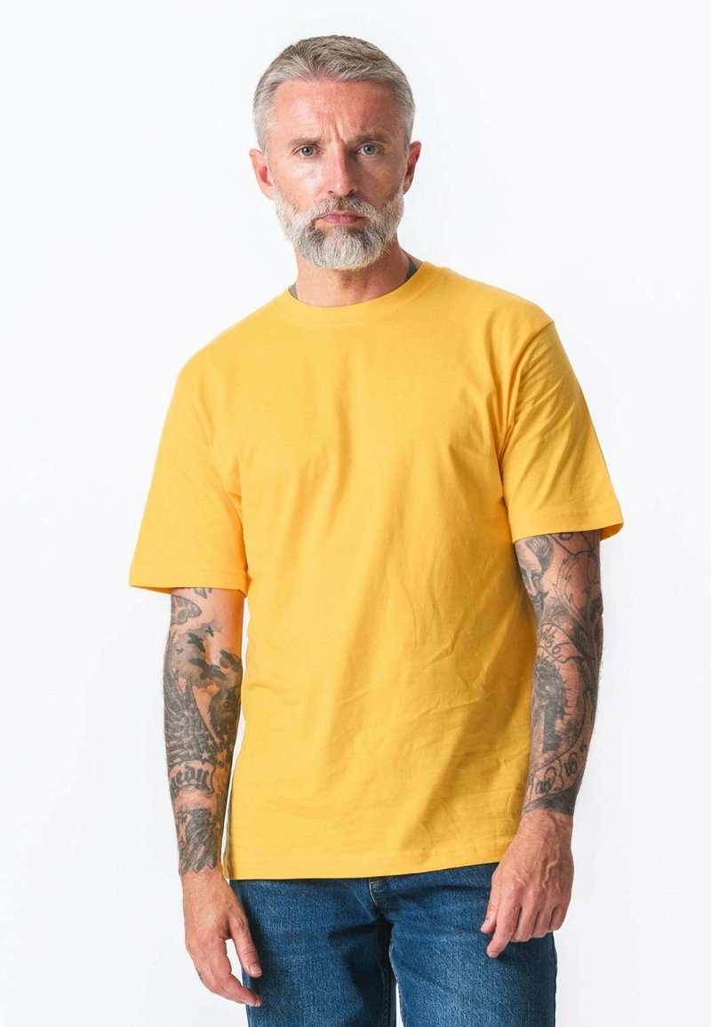 Middelaldrende mand med grå skæg og tatoveringer på begge arme iført en gul kortærmet skjorte og blå jeans, stående foran en hvid baggrund.
