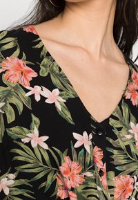 Schwarzer Stoff mit einem lebhaften Blumenmuster aus rosa und weißen Blumen. V-Ausschnitt mit Knöpfen vorne. Eingewebte strukturierte Blätter.
