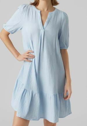Donna che indossa un vestito azzurro chiaro, ampio, con maniche corte a sbuffo e orlo arricciato su uno sfondo semplice.