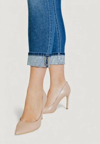 Beiget, korkokengät, joissa on teräväkärkinen muotoilu. Mukana siniset farkut, joissa on kalvosin, joka on koristeltu kimaltelevilla yksityiskohdilla.