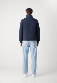 Μπλέ ναυτικού πηλίκιο μπουφάν με ψηλό γιακά, ραβδωτούς καρπούς και λεία υφή, συνδυασμένο με ανοιχτό μπλε τζιν και λευκά αθλητικά παπούτσια.