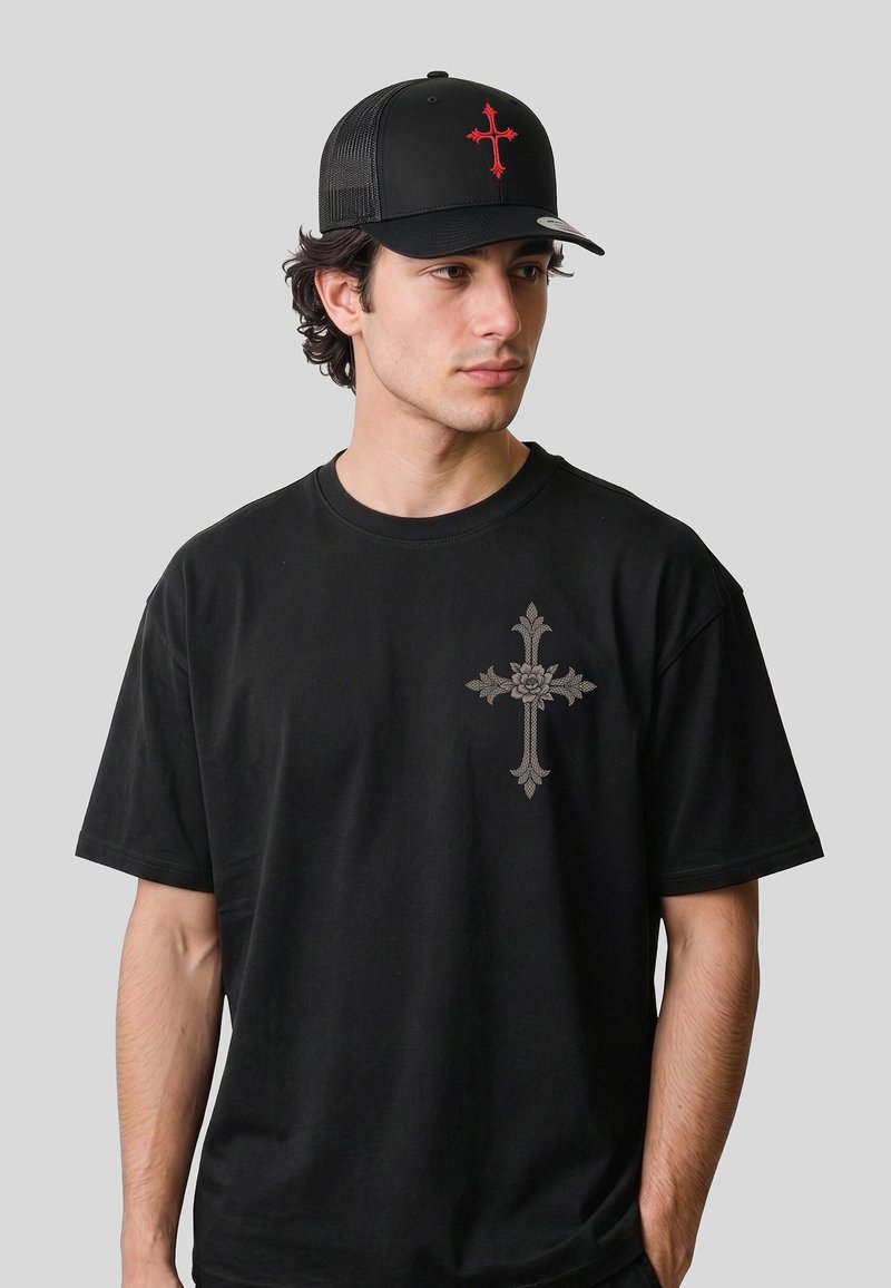 Jeune homme portant un T-shirt noir avec un motif de croix florale grise et une casquette en filet noire avec un emblème de croix rouge, regardant sur le côté.