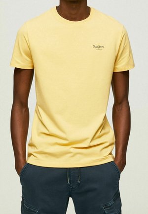 Mann trägt ein schlichtes gelbes kurzärmeliges T-Shirt mit kleinem Logo auf der Brust und dunkelblaue legere Hose mit Händen in den Taschen.