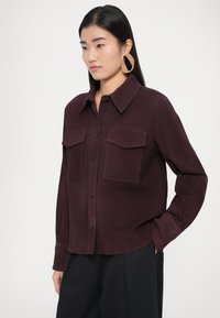 Cămașă burgundy cu nasturi, două buzunare la piept, nasturi negri și guler clasic, asortată cu pantaloni negri și cercei îndrăzneți aurii.