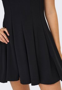 Jupe plissée noire portée par une personne avec une bague à la main gauche, montrée du milieu de la cuisse à la taille sur un fond uni.