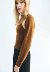 Blonde Frau, die einen braunen asymmetrischen Pullover mit einem langen Ärmel und schwarzen Hosen trägt, steht vor einem schlichten hellen Hintergrund.