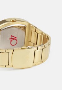 Goldfarbene Uhr mit rechteckigem Zifferblatt, Metallarmband und Verschluss. Die Rückseite zeigt eine silberne Oberfläche mit der roten Zahl "40" und dem Markennamen.