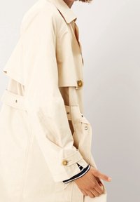 Gabardina beige con cintura ceñida, diseño cruzado con doble botonadura y detalles con botones. Cuenta con cuello y mangas entalladas. Textura suave.