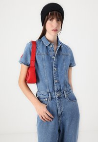 Denim overal s krátkymi rukávmi, modrá látka, zapínanie na gombíky vpredu a dve predné vrecká. V kombinácii s červenou koženou taškou a čiernym pleteným čiapkom.