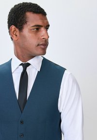 Tangerfarbenes, strukturiertes Weste mit tiefem V-Ausschnitt, ausgestattet mit zwei Knöpfen, getragen über einem weißen Hemd und ergänzt durch eine schwarze Krawatte.