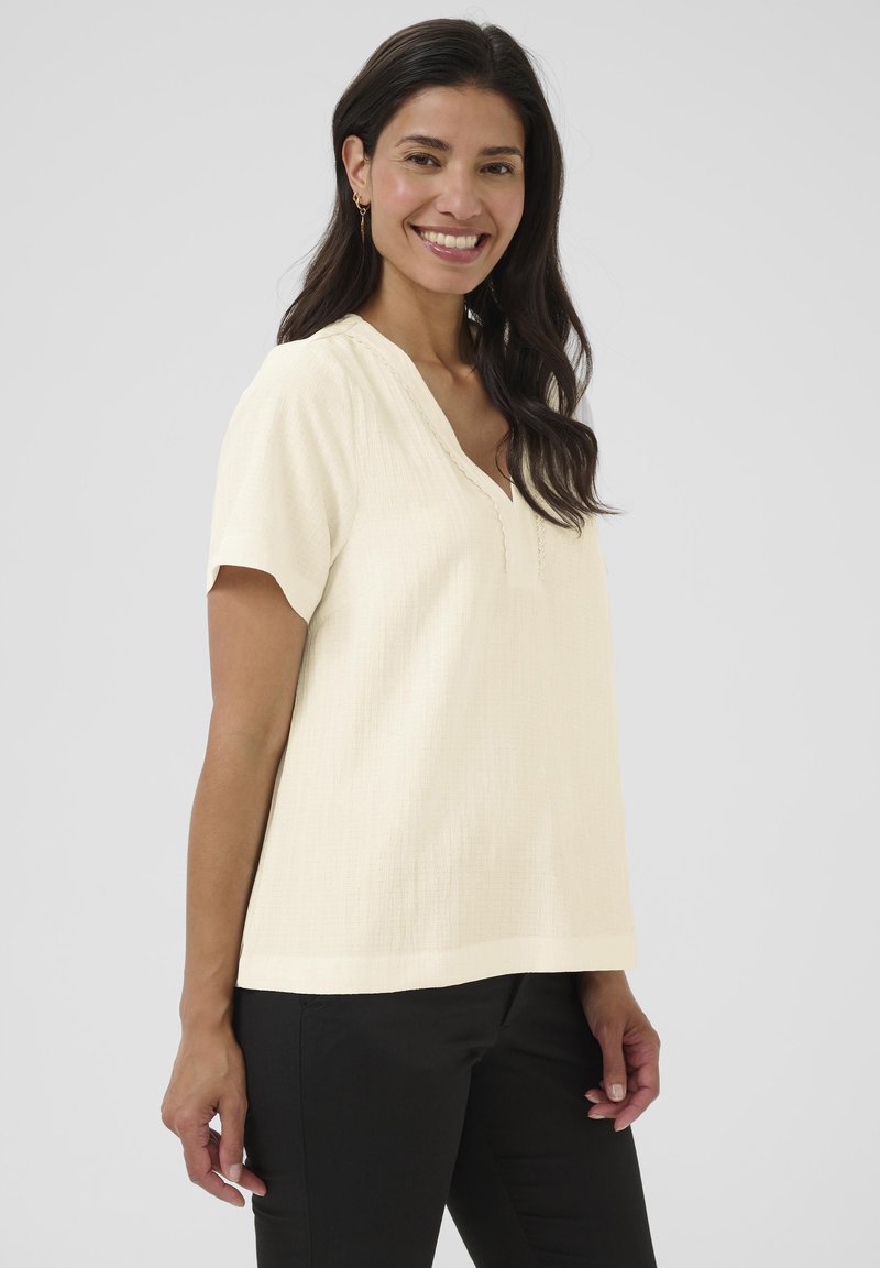Blouse courte à manches courtes de couleur beige clair avec un col en V et un tissu texturé, présentant des accents festonnés le long de l'encolure et une coupe décontractée.
