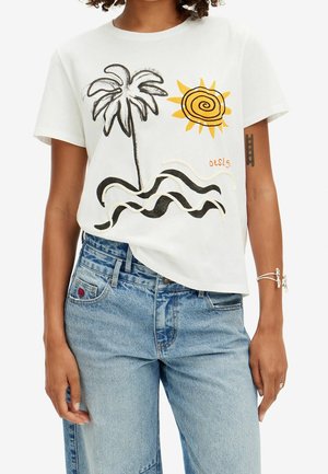 Person wearing a white t-shirt with a black palm tree, yellow sun, wavy black lines, and blue jeans, showing a tattoo and silver bracelets.