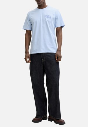 Homme portant un t-shirt bleu clair avec l'inscription « The Shadow Depth », un jean large foncé et des chaussures en cuir marron, debout sur fond blanc.