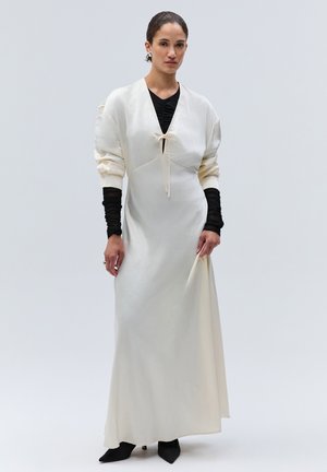 Donna che indossa un lungo vestito bianco con un nodo frontale sopra una maglia nera a maniche lunghe e scarpe nere con punta, in piedi su uno sfondo semplice.