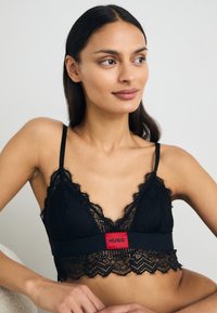 Svart blonde bralette med dyp utringning, delikate bølgede kanter og et bredt rødt logobånd nederst. Justerbare stropper.