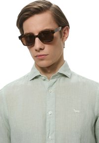 Occhiali da sole tartarugati marroni con lenti scure, indossati con una camicia button-up in lino verde chiaro con un piccolo logo ricamato.