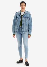 Chaqueta de mezclilla azul claro con diseño clásico, cierre de botones, dos bolsillos en el pecho, usada sobre un suéter de punto verde y jeans ajustados azul claro. Mocasines negros.