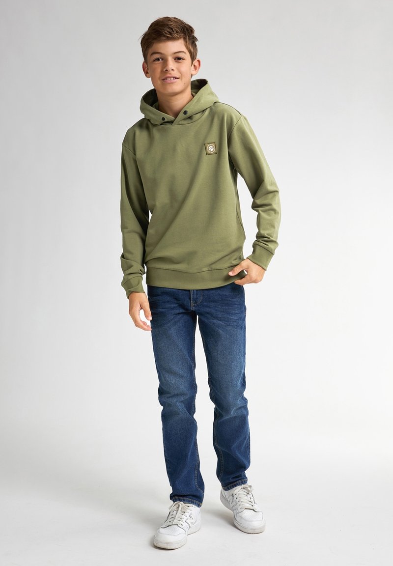 Felpa verde con cappuccio regolabile e una piccola toppa con logo, abbinata a jeans blu e sneakers bianche su uno sfondo chiaro.