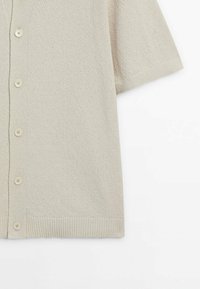Vaaleanbeige lyhythihainen kardigan, jossa on tekstuuri-neulekuvio, painonappietu ja ribbaushelman yksityiskohdat.