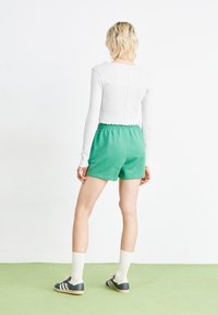 Πράσινες βαμβακερές βραχιώνες με ελαστική μέση, συνδυασμένες με ένα λευκό μακρυμάνικο μπλουζάκι, μαύρα παπούτσια και λευκές κάλτσες. Εμφανίζεται πίσω όψη.