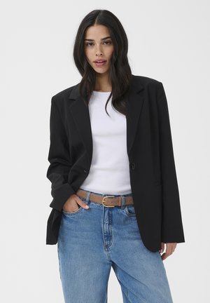 Femme aux longs cheveux foncés portant un haut blanc, un blazer noir surdimensionné, un jean bleu et une ceinture marron, debout avec une main dans la poche.