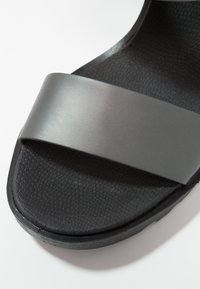 Sandale noire avec une bride grise mate. La semelle intérieure texturée présente un motif en relief, et la semelle extérieure est entièrement noire et légèrement incurvée.