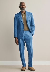 Blauwe linnen pak met een notch lapel en twee knopen, gecombineerd met een bruine geribde polo. Braune schoenen maken de look compleet.