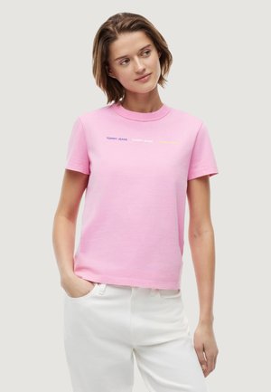 Frau trägt ein pinkes Tommy Jeans T-Shirt und weiße Hose, steht mit einer Hand in der Tasche und schaut leicht zur Seite.