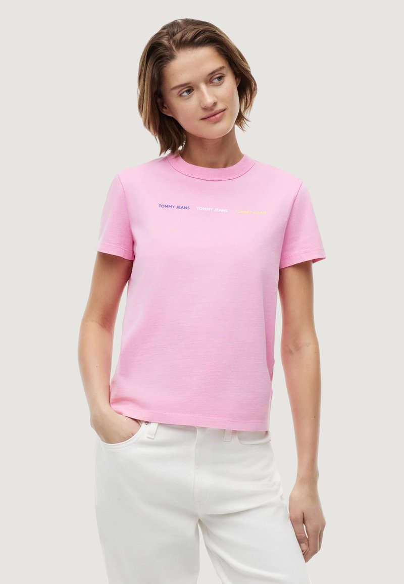 Femme portant un t-shirt rose Tommy Jeans et un pantalon blanc, debout avec une main dans la poche, regardant légèrement sur le côté.