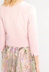 Giacca corta rosa chiaro con maniche a tre quarti, abbinata a una gonna trasparente ricamata con motivi floreali multicolori e texture variegate.