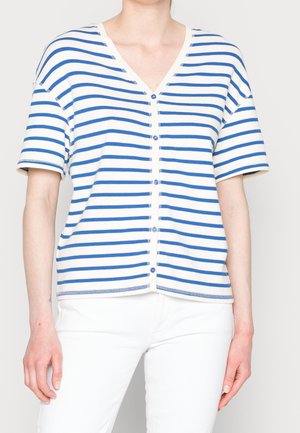 Personne portant une chemise à manches courtes boutonnée à rayures horizontales blanches et bleues avec un pantalon blanc, debout devant un fond uni.