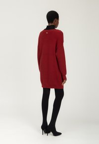 Rode gebreide oversized trui met lange mouwen, geribbelde textuur, minimale details aan de achterkant, gecombineerd met zwarte panty en hoge hakken.