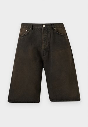Mężczyzna ubrany w luźną różową bluzę z zamkiem, czarny t-shirt, ciemne szorty do kolan, czarne skarpetki i czarne sznurowane buty na jednolitym tle.