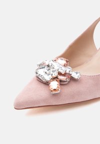 Ροζ σουέτ γόβα με μυτερή μύτη και ψηλό τακούνι, με συγκεντρωμένες διαφανείς και ροδακινί πολύτιμες πέτρες στο μπροστινό μέρος.