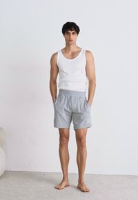 Grå treningsshorts med elastisk midje og sidelommer, kombinert med en hvit tanktopp. Modellen står barbeint mot en enkel bakgrunn.