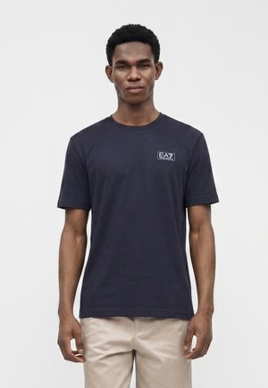 Homme portant un t-shirt noir uni à manches courtes avec un petit logo EA7 sur la poitrine et un pantalon beige, debout devant un fond blanc uni.