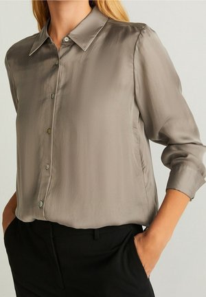 Femme portant une chemise grise en satin à manches longues boutonnée, rentrée dans un pantalon noir, les mains dans les poches.