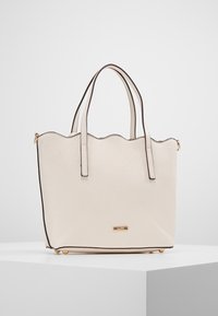 Beige synthetic tote bag with scalloped top edge, black trim, dual handles, gold logo plate, and four gold-tone feet at the base.