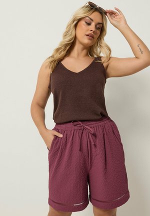 Kvinde med blondt hår iført brun ærmeløs top og teksturerede lilla shorts, holder solbriller over hovedet, hånd i lommen.